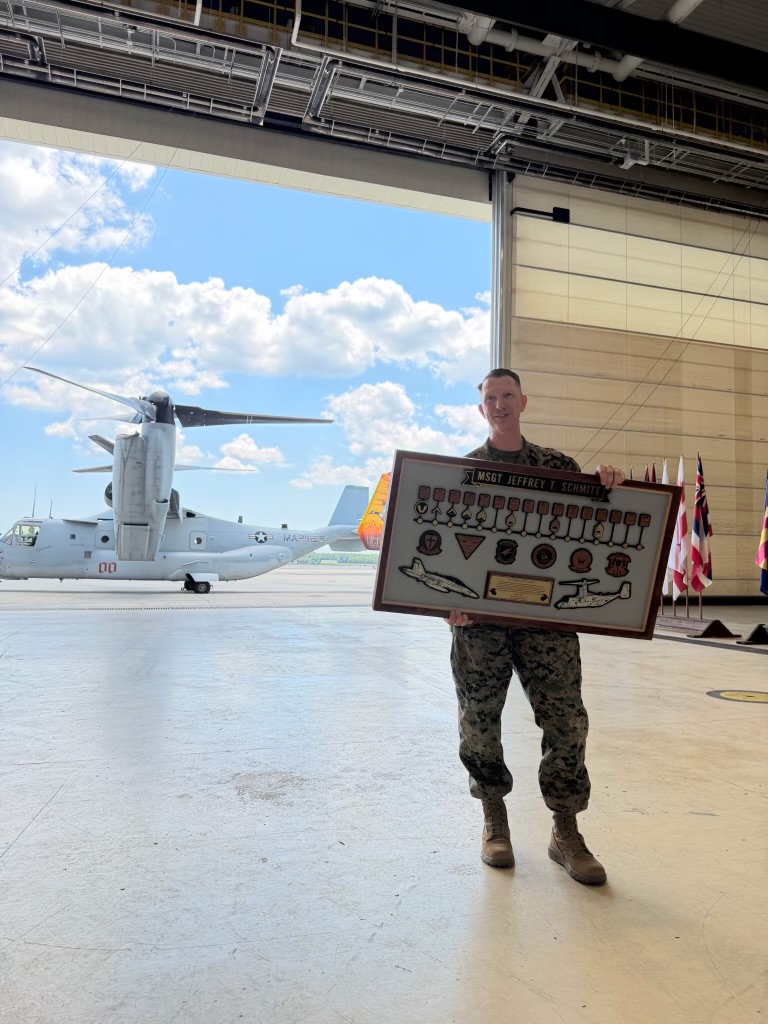 MSgt Jeffrey Schmitt holding his career timeline plaque beside an MV-22 Osprey